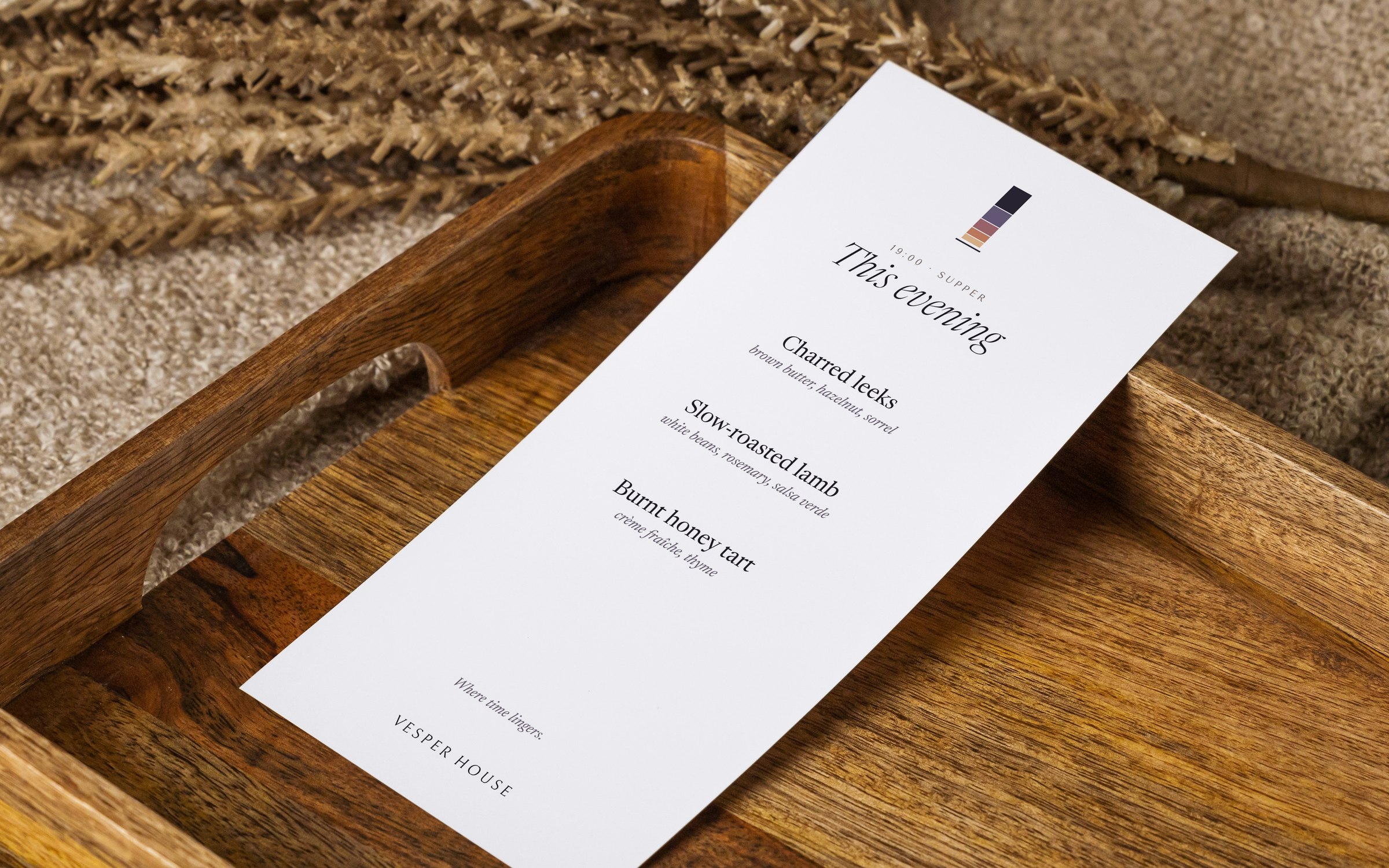 A tall Vesper House supper menu titled This evening, resting on a wooden tray beside dried grasses and linen.