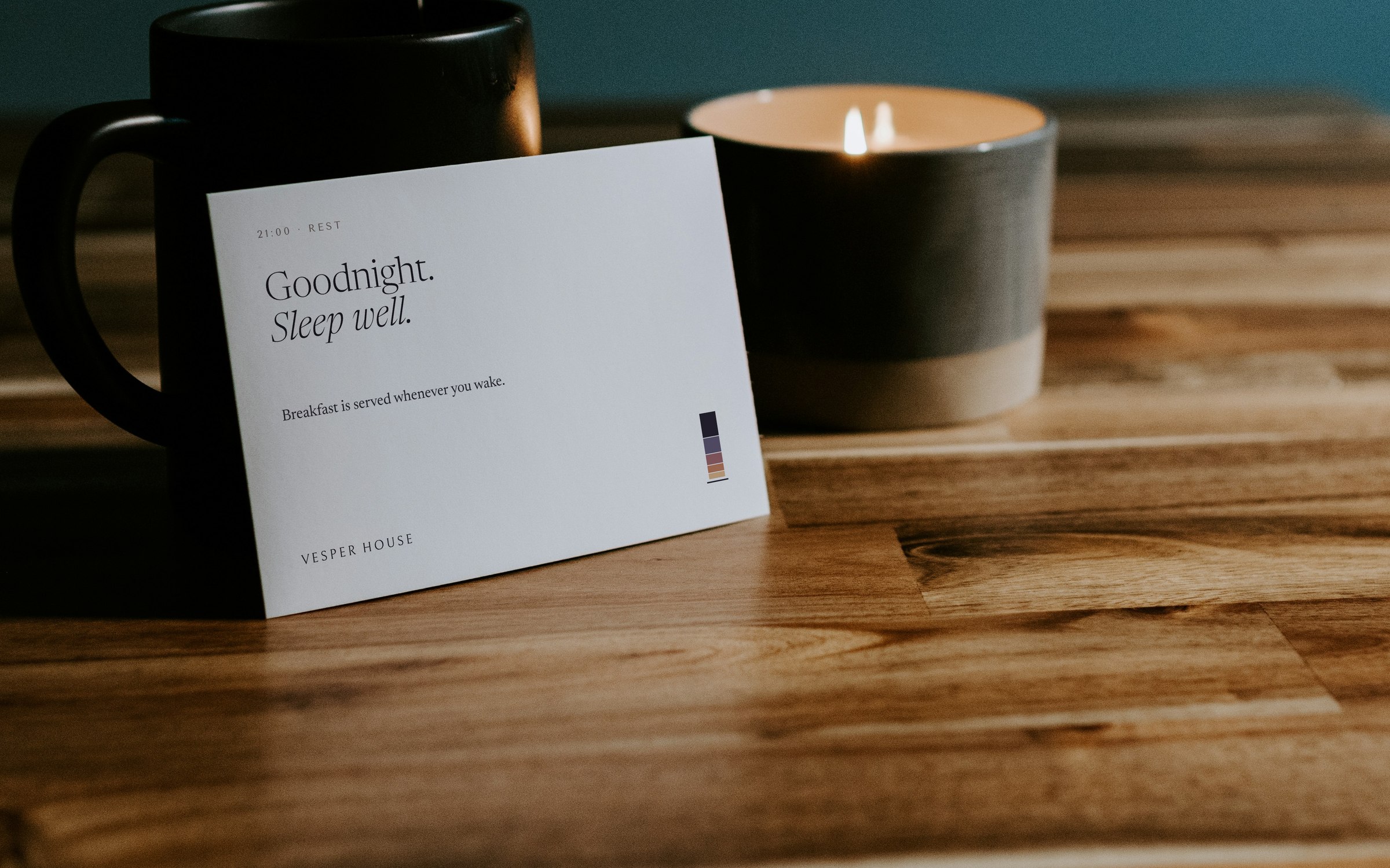 A Vesper House turndown card reading Goodnight. Sleep well., propped against a mug beside a lit candle on a wooden table.