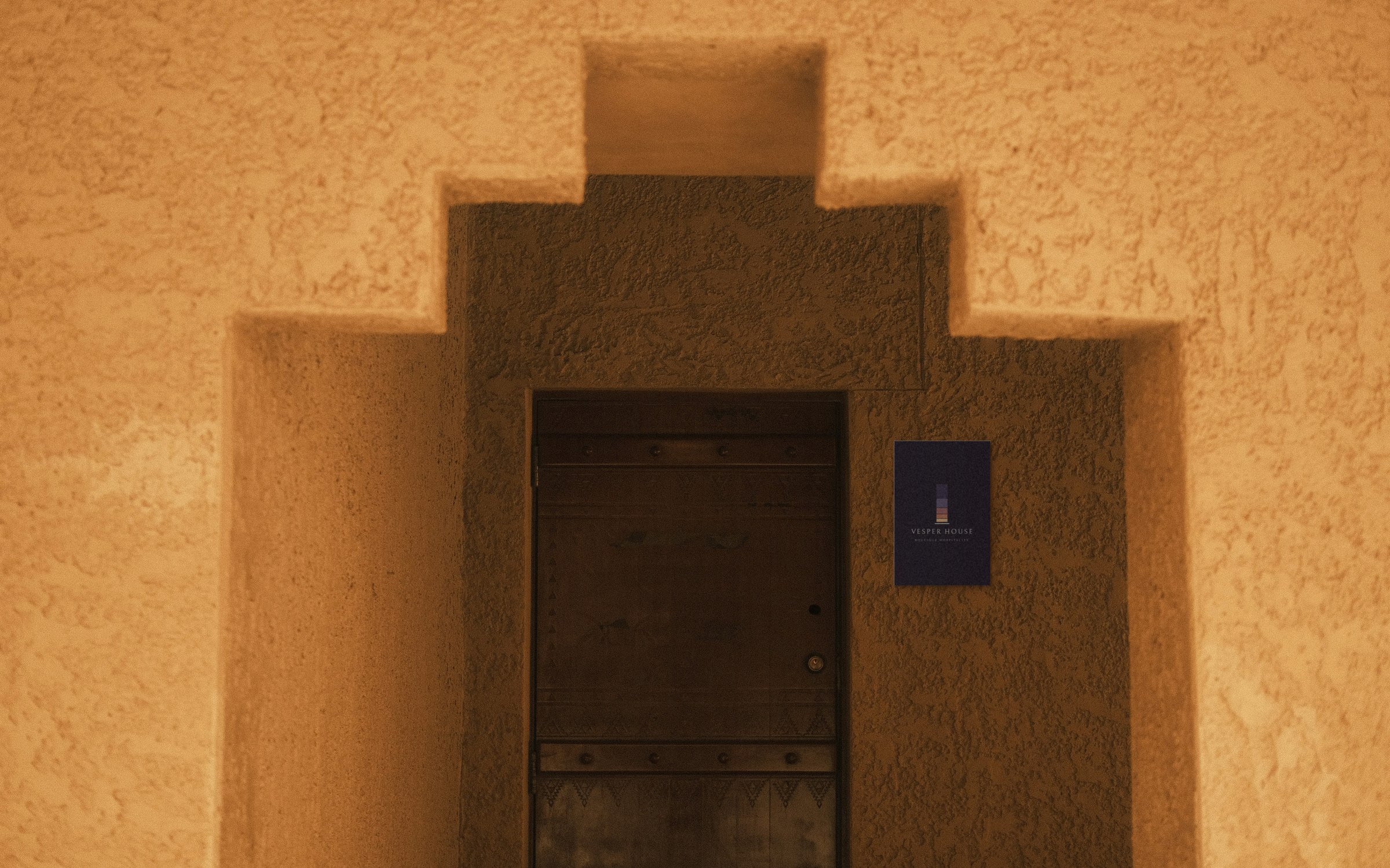 A blackened steel Vesper House plaque, with the window symbol and Boutique Hospitality, mounted beside a deep doorway in warm textured plaster.
